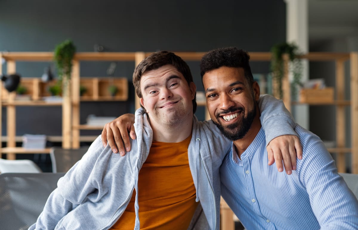 Two smiling men sit closely together, one with his arm around the other, showing a warm and friendly bond in a modern indoor setting.